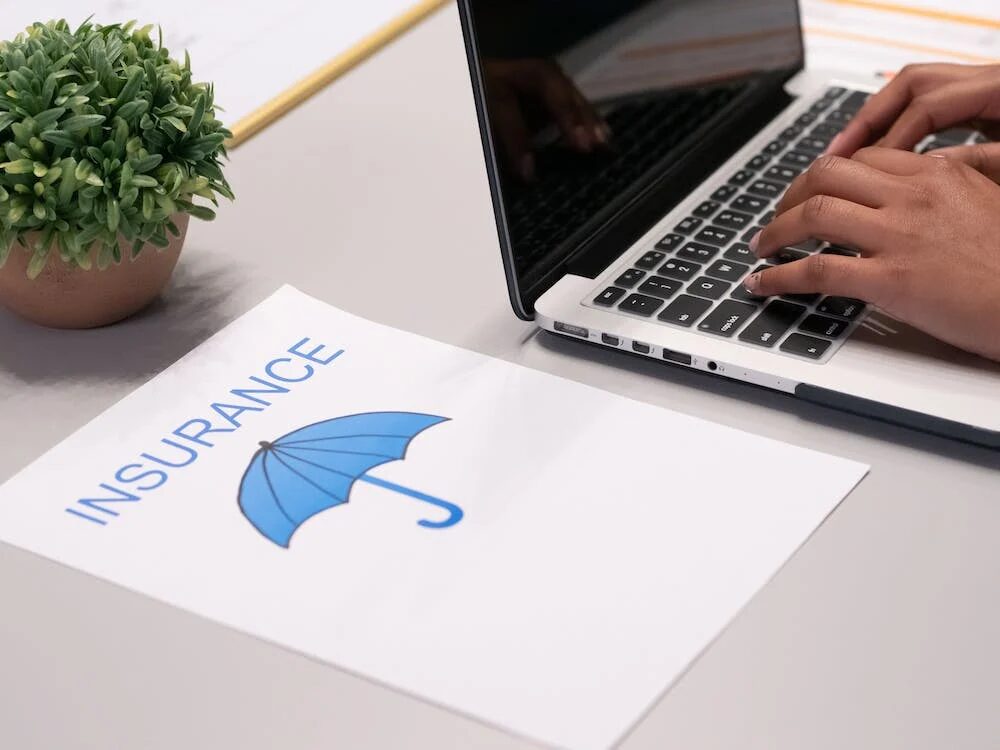 a blue umbrella printed on a paper with the word 'insurance' beside a person working on a laptop