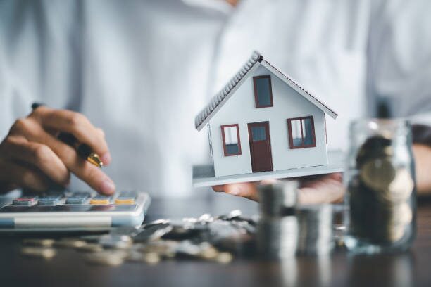 hands of a person using the calculator while holding a house model on another hand with coins on the table