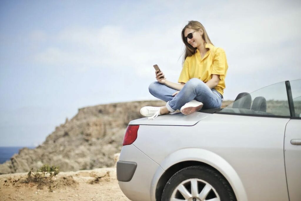 a woman happily sitting on the trunk of her car while looking at her phone